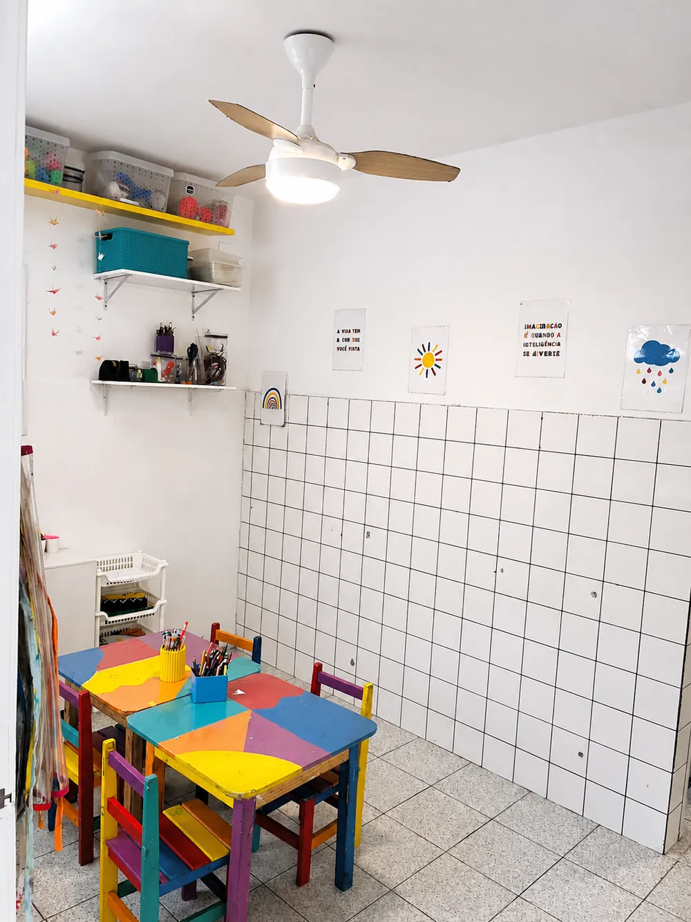 Ateliê com mesas coloridas, potes de lápis, prateleiras de materiais e cartazes na parede.
