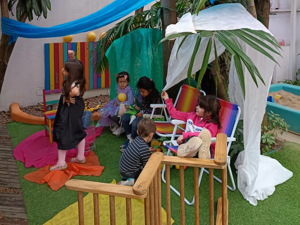 Crianças brincando livremente no quintal, entre cabanas improvisadas com tecidos, cadeiras e tecidos coloridos no chão.