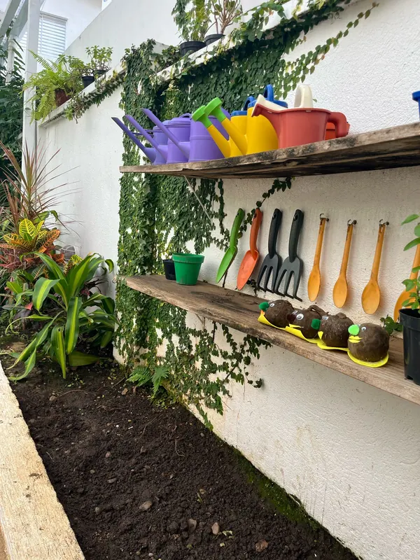 Canteiro de jardinagem com terra preparada, regadores coloridos e ferramentas penduradas nas prateleiras.