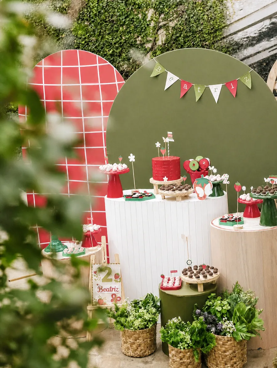Mesa de festa com tema de morango: painéis verde e vermelho, bolo, docinhos e vasos de plantas.