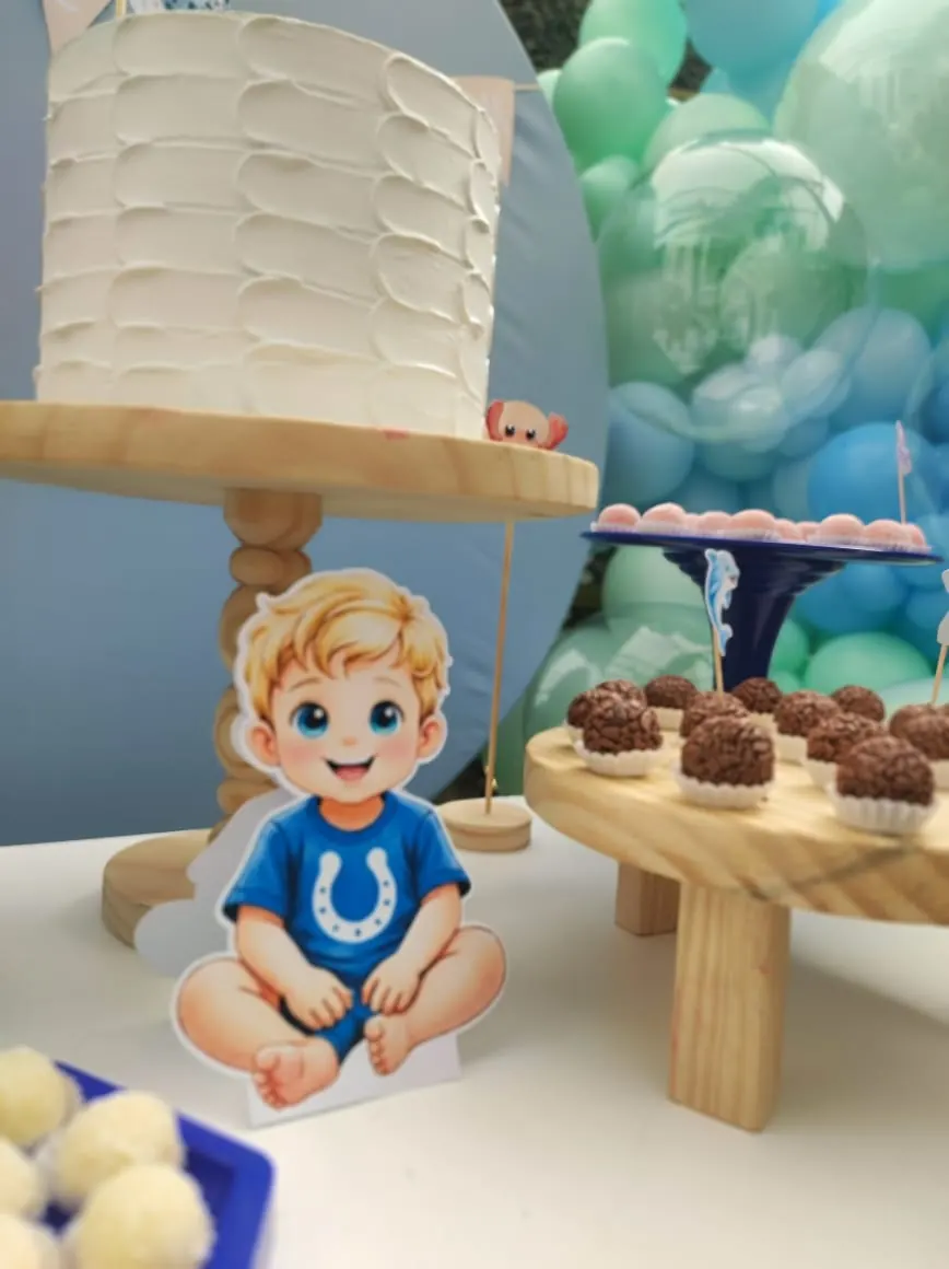 Mesa em tons de azul com balões, bolo branco, docinhos e recorte do aniversariante.