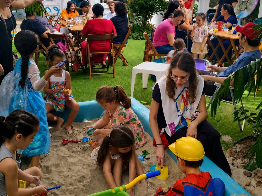 Brincante agachada no tanque de areia brincando com as crianças durante uma festa, com as famílias às mesas ao fundo.