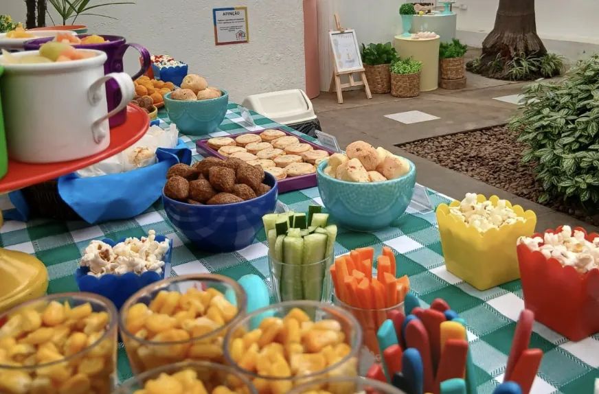 Mesa de festa montada no quintal, com tigelas coloridas de salgados, frutas, legumes e pipoca sobre toalha xadrez.