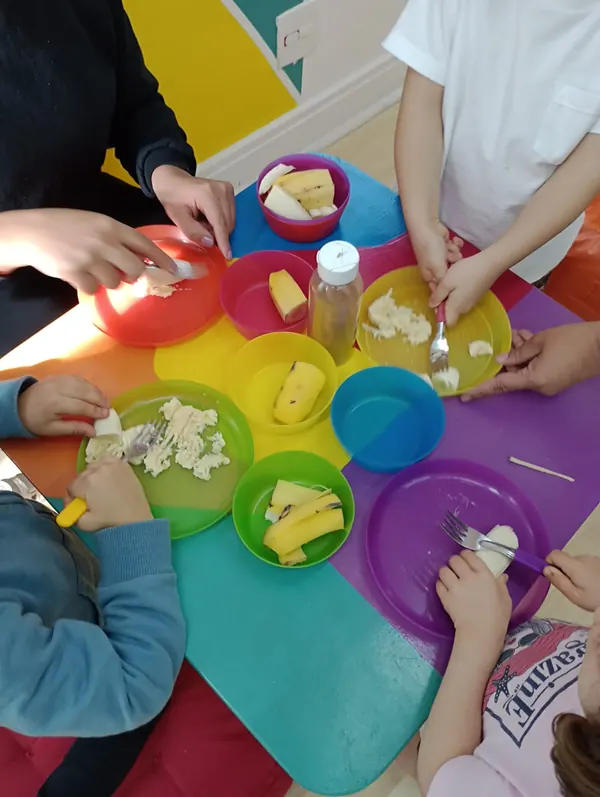 Crianças amassando banana com garfos em pratinhos coloridos durante a oficina de culinária.