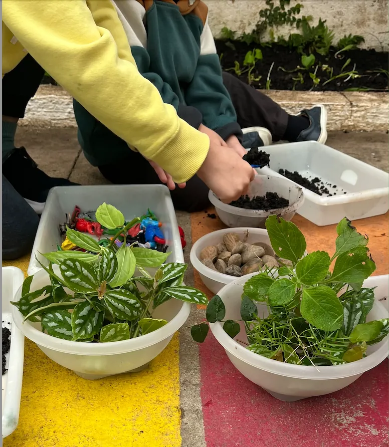 Crianças manuseando terra com pá enquanto plantam mudas em bacias, durante a oficina de jardinagem.