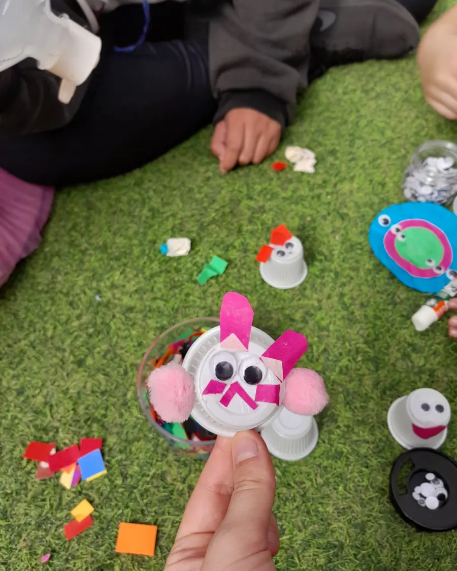 Bichinhos criados com tampinhas, olhos móveis, pompons e papel colorido sobre a grama.