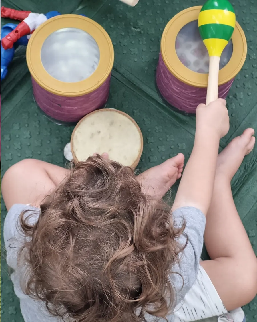 Criança pequena sacudindo um chocalho ao lado de tambores e pandeiro no chão.