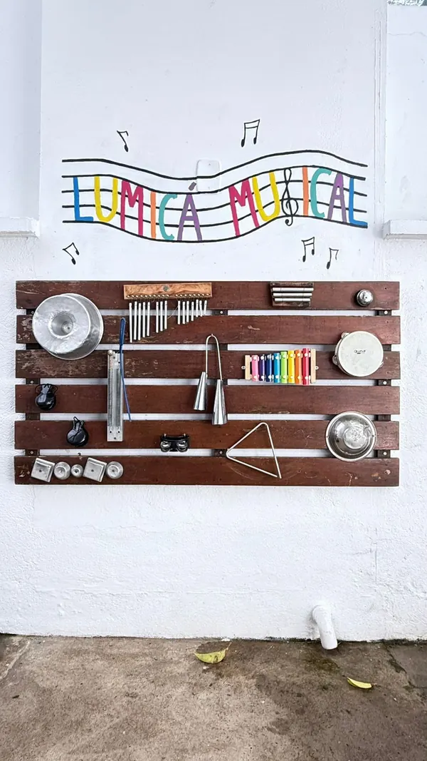 Painel musical de madeira no quintal, com xilofone, triângulo, pandeiro e outros instrumentos, sob o letreiro pintado Lumicá Musical.