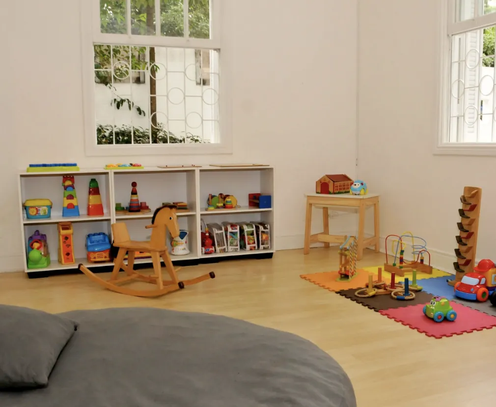 Sala baby com estantes de brinquedos, cavalinho de madeira, tapete emborrachado e colchonete no chão.