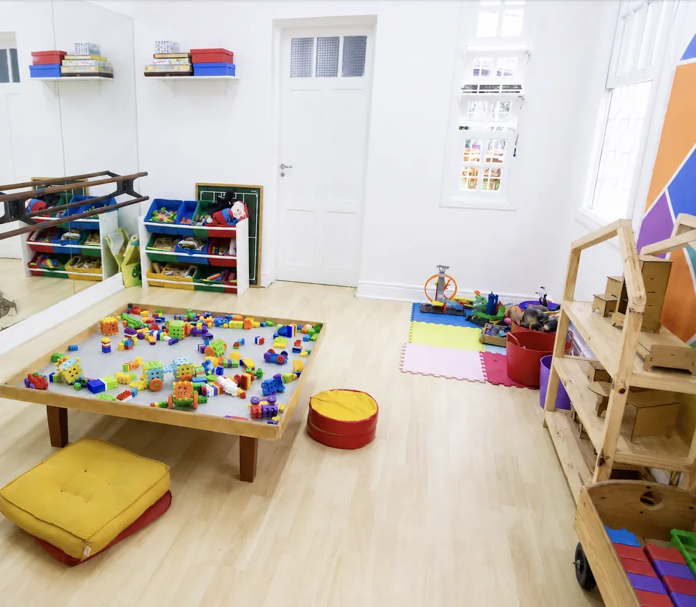 Sala kids com mesa baixa de blocos, almofadas de chão, prateleiras de brinquedos e barra de espelho.