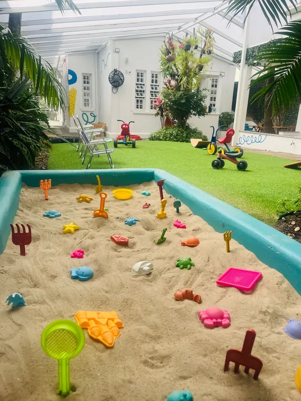 Tanque de areia com formas e pás coloridas, com o quintal e os triciclos ao fundo.