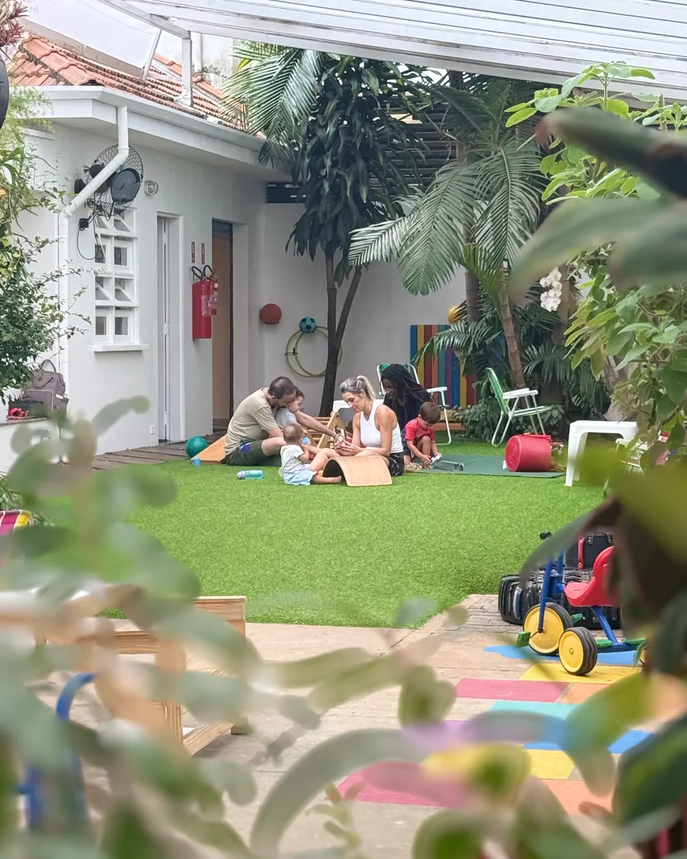 Famílias e crianças sentadas na grama do quintal coberto, entre plantas e brinquedos.