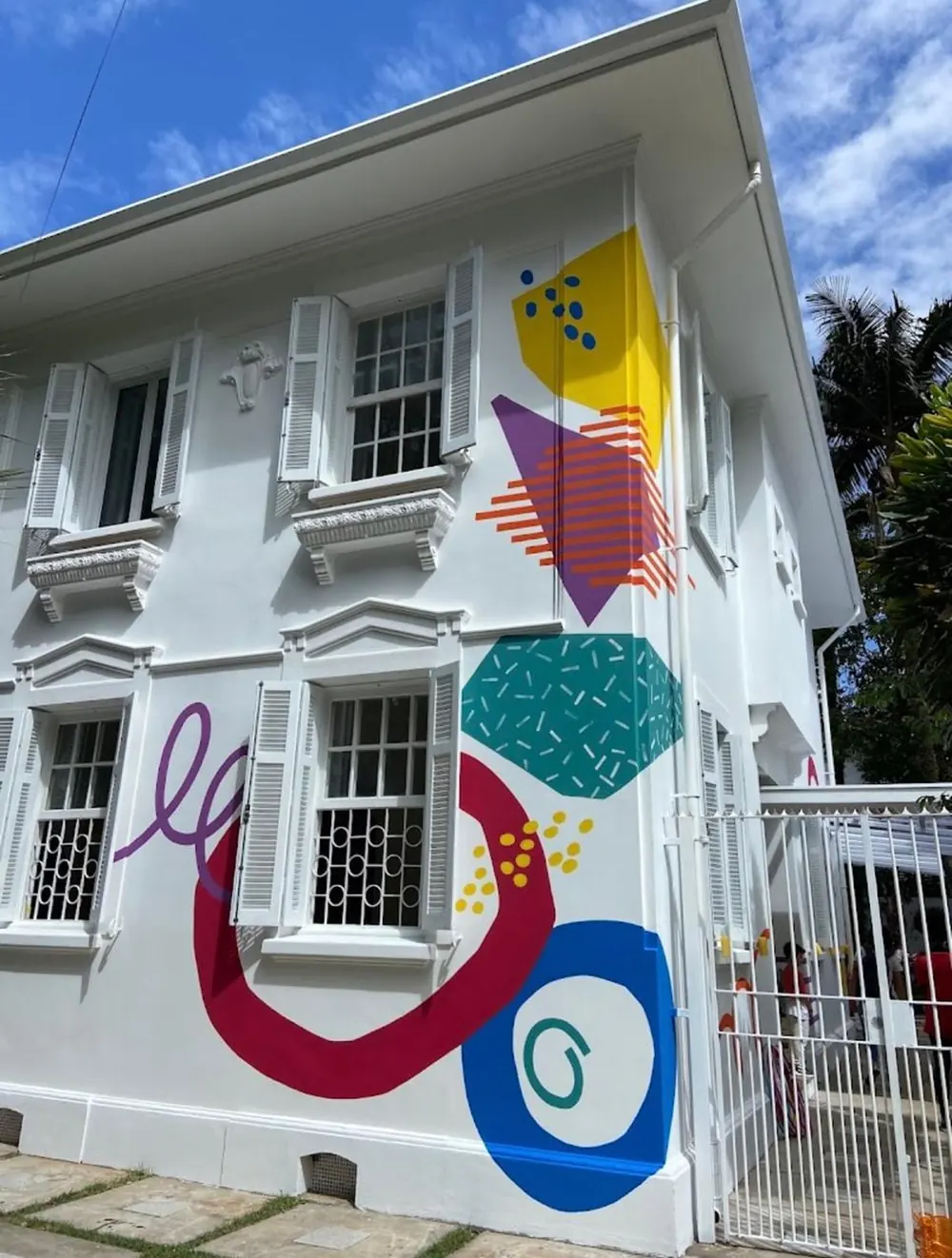 Fachada branca da Lumicá com o mural colorido, sob céu azul.